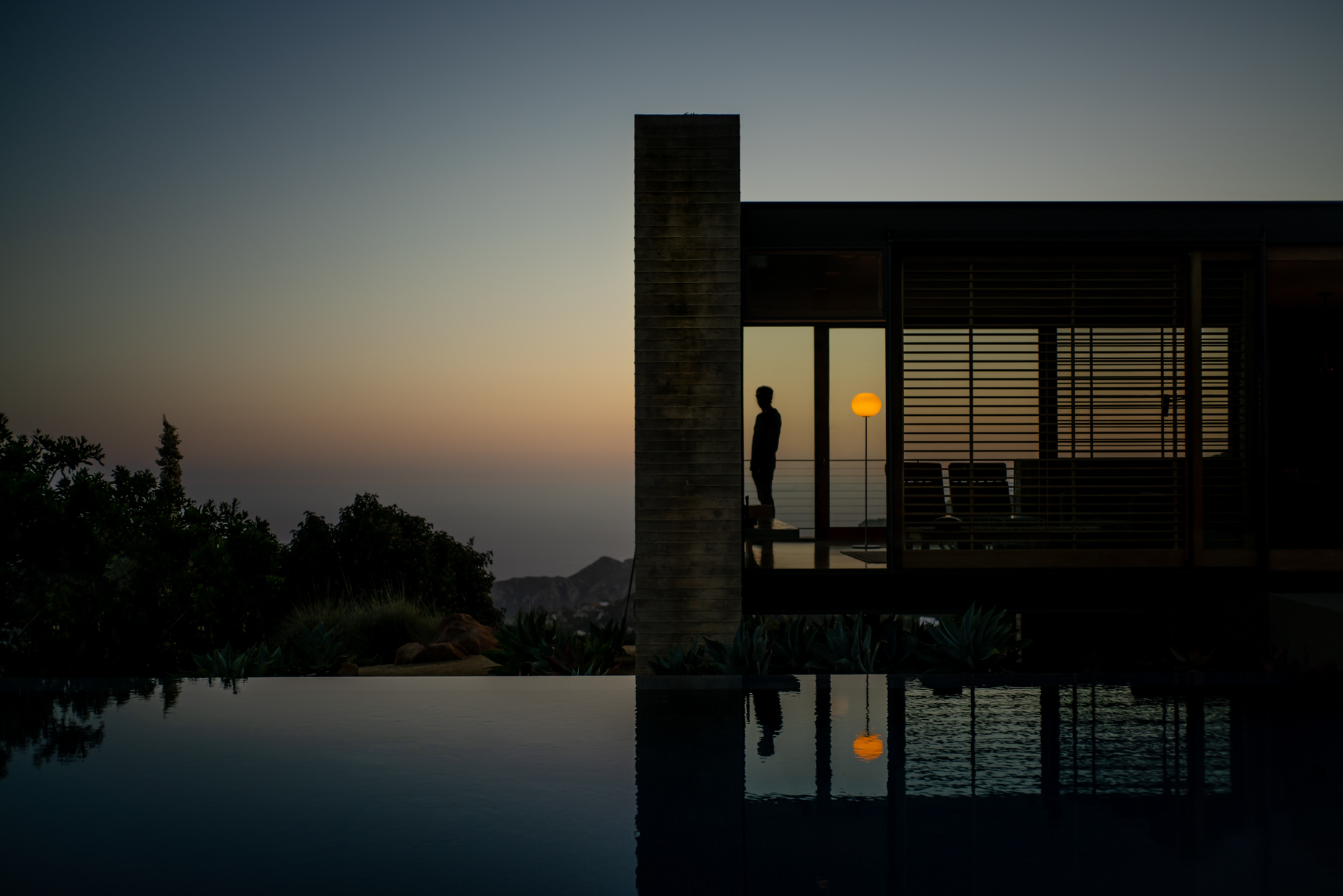 Man standing in modern house looking out over ocean and infinity pool during sunset