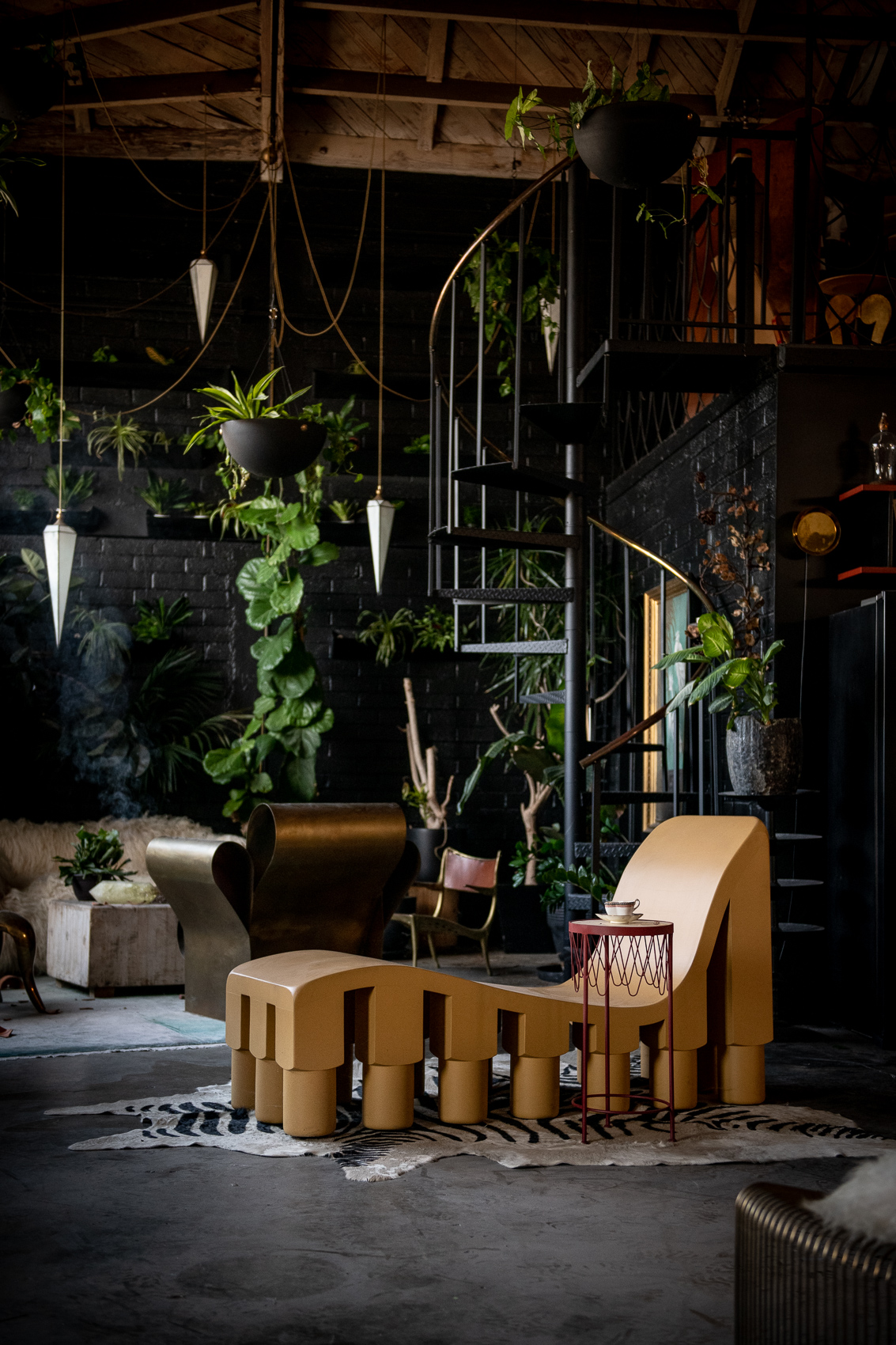 Yellow chaise lounge on zebra skin in front of black circular staircase and plants.