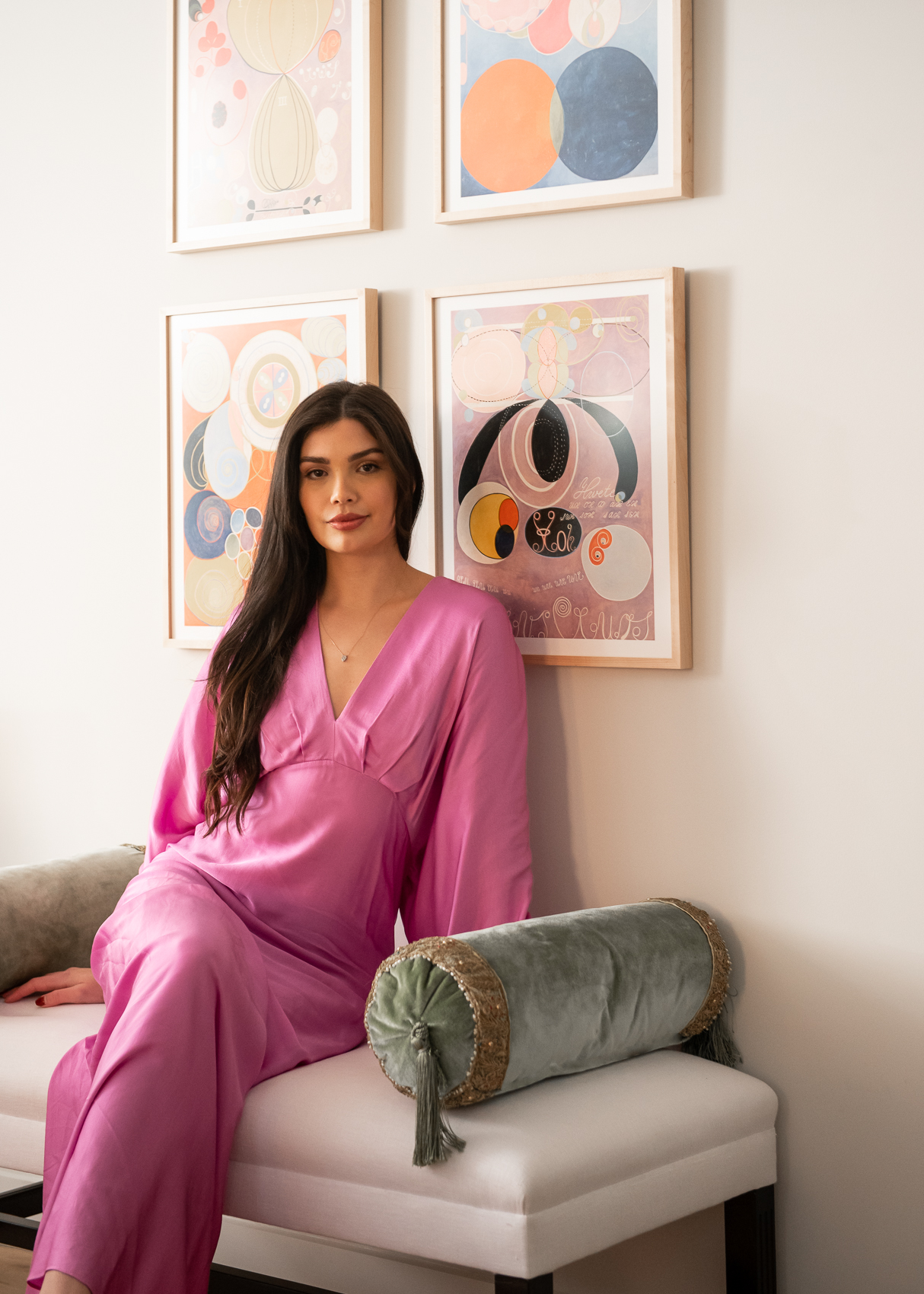 Woman in pink jumpsuit sits on plush day bed under four framed wall prints